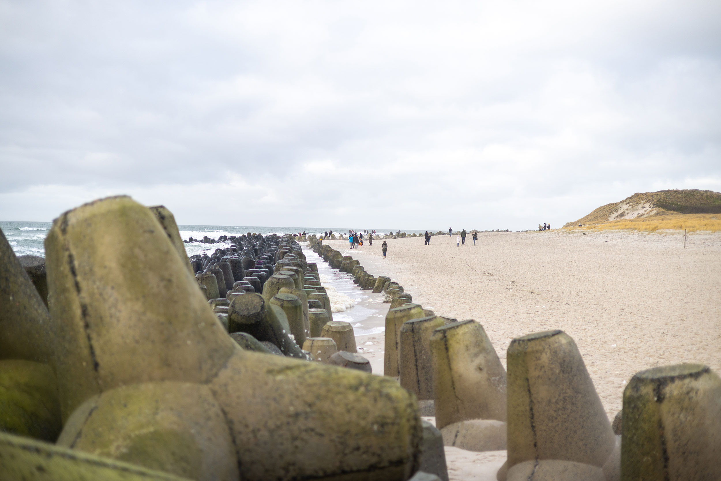 Große Wellenbrecher aus Beton säumen den Rand eines Sandstrandes, während in der Ferne Menschen unter einem bewölkten Himmel spazieren gehen.