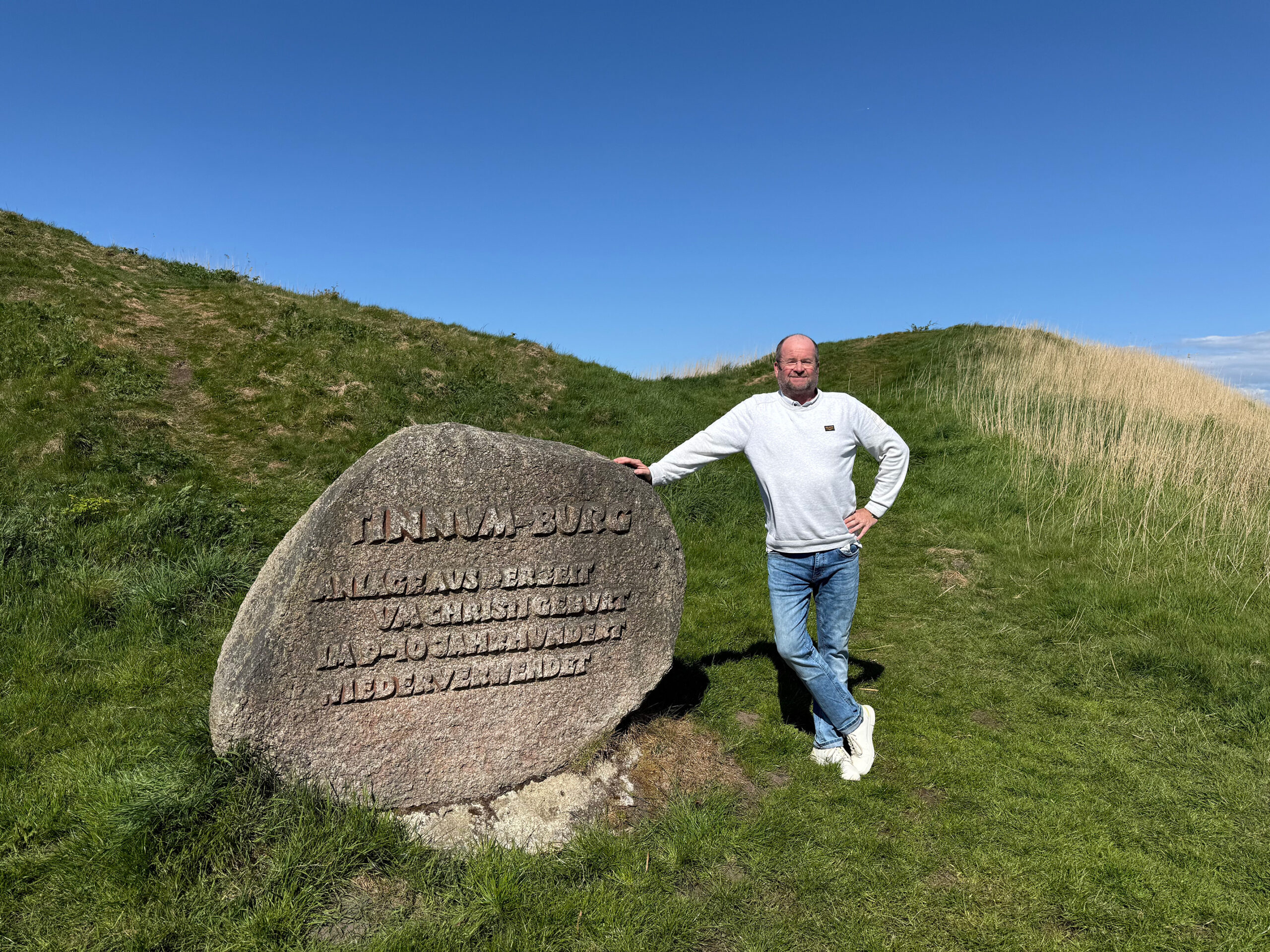 Ein Mann steht unter freiem Himmel neben einem großen eingemeißelten Stein auf einem grasbewachsenen Hügel unter blauem Himmel.