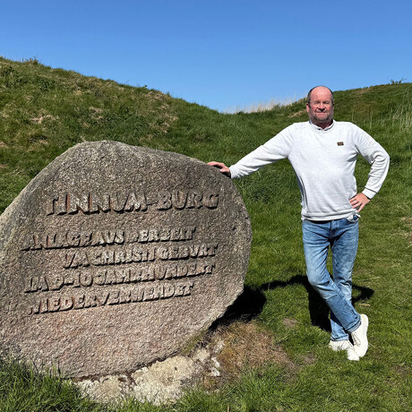 Ein Mann in weißem Pullover und Jeans steht neben einem großen eingravierten Stein auf einem grasbewachsenen Hügel unter einem klaren blauen Himmel.