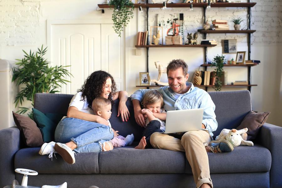 Junge Familie sitzt lachend auf der Couch mit Laptop auf dem Schoß.
