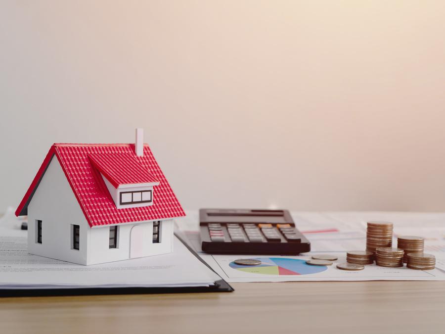 Modellhaus neben Taschenrechner, Finanzunterlagen und Geldstapel.