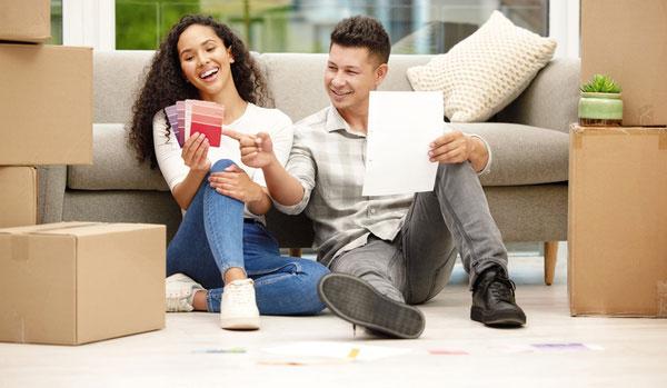Junges lachendes Paar sitzt vor Couch in neuer Wohnung mit Umzugskartons.