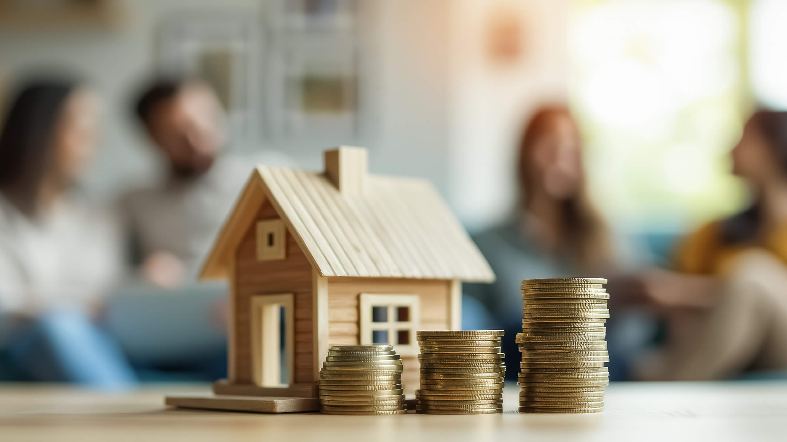 Holzhaus und Münzen auf Tisch, vier Personen im Hintergrund.