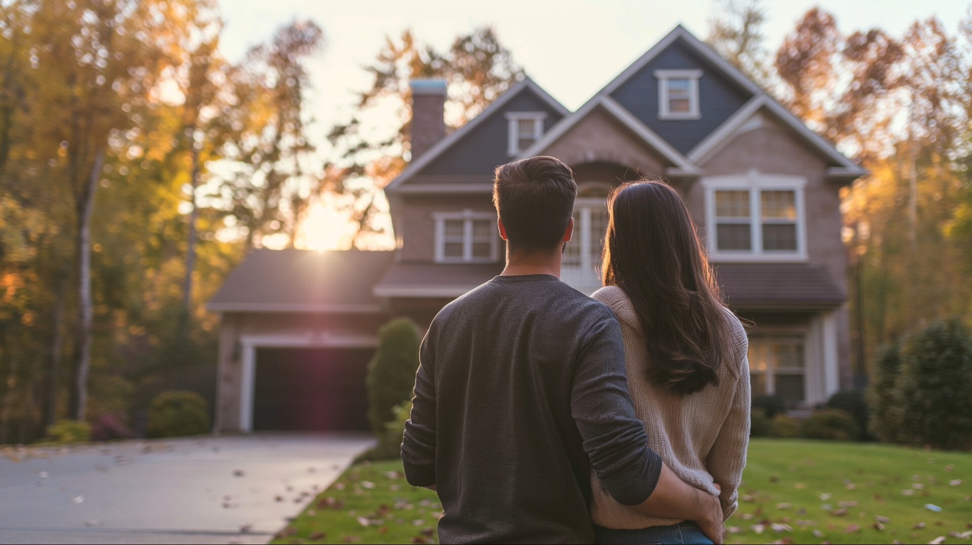 Ein junges Paar blickt von hinten auf ein großes Einfamilienhaus im Sonnenuntergang.