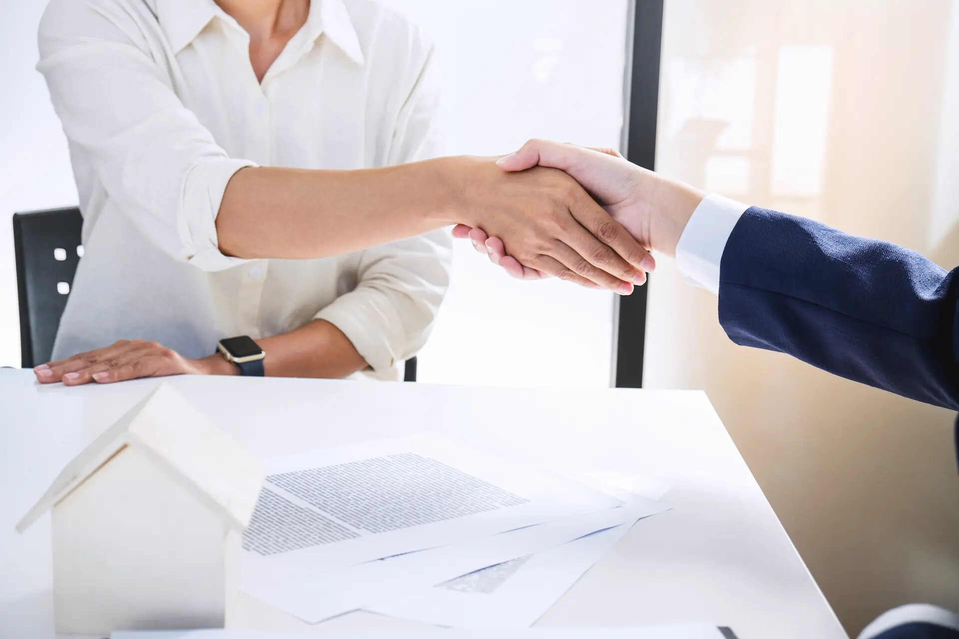 Händedruck über einen Schreibtisch mit Papieren und Hausmodell.
