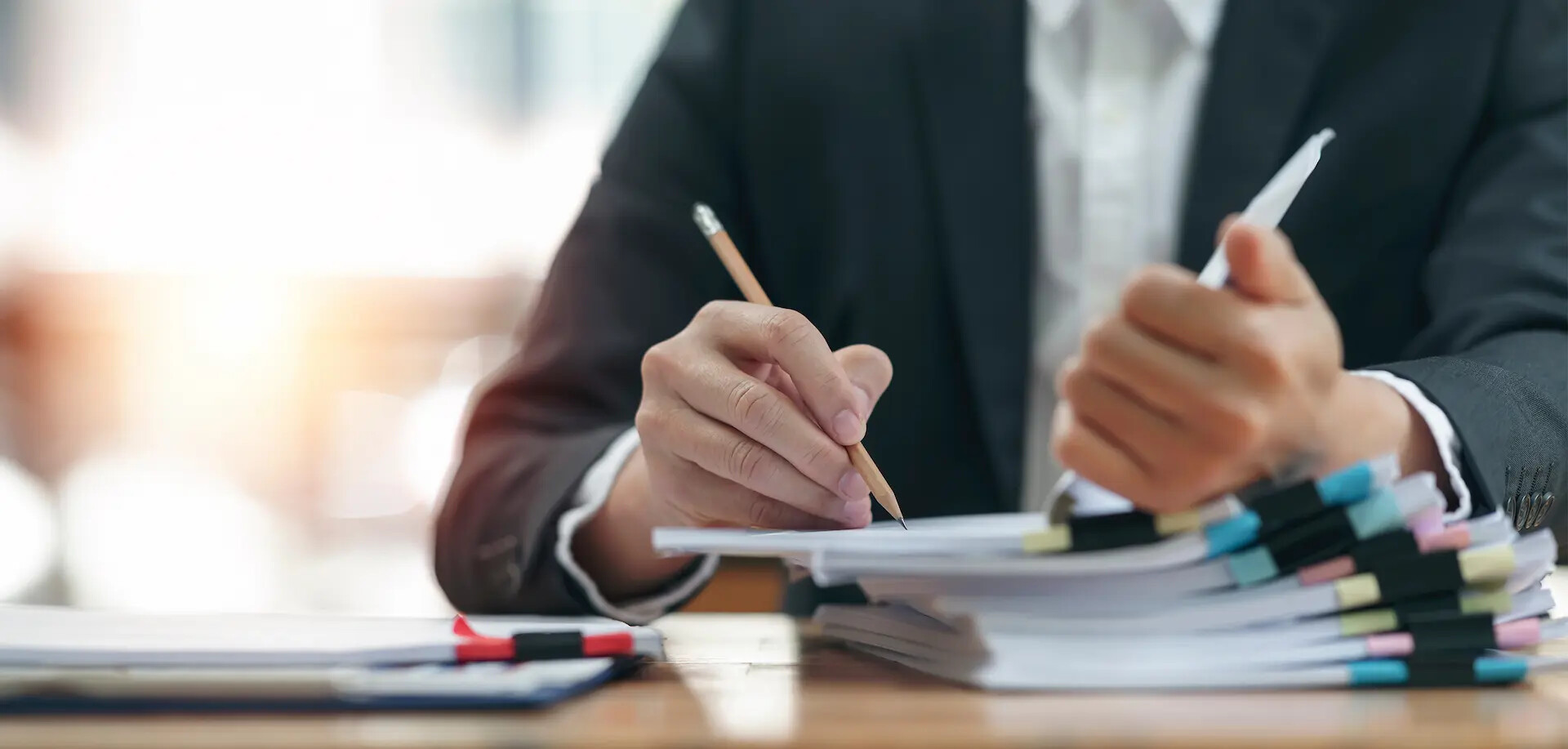 Person im Anzug prüft Dokumente an Schreibtisch mit Papierstapeln.