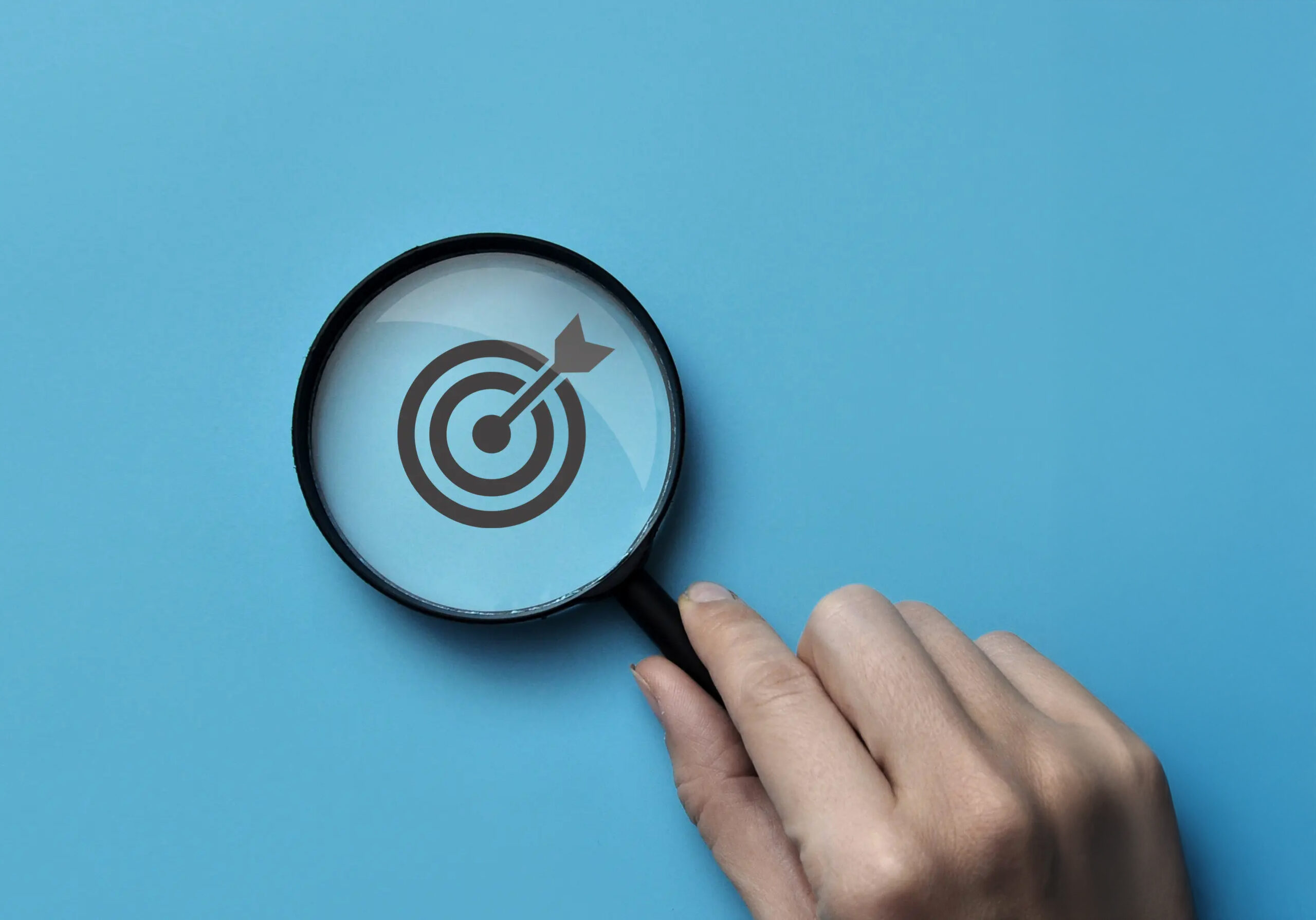 Hand mit Lupe über Zielscheibe mit Pfeil auf blauem Hintergrund.