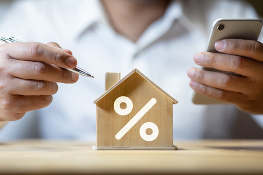 Kleines Modellhaus mit Prozentzeichen, Mensch mit Stift und Handy dahinter