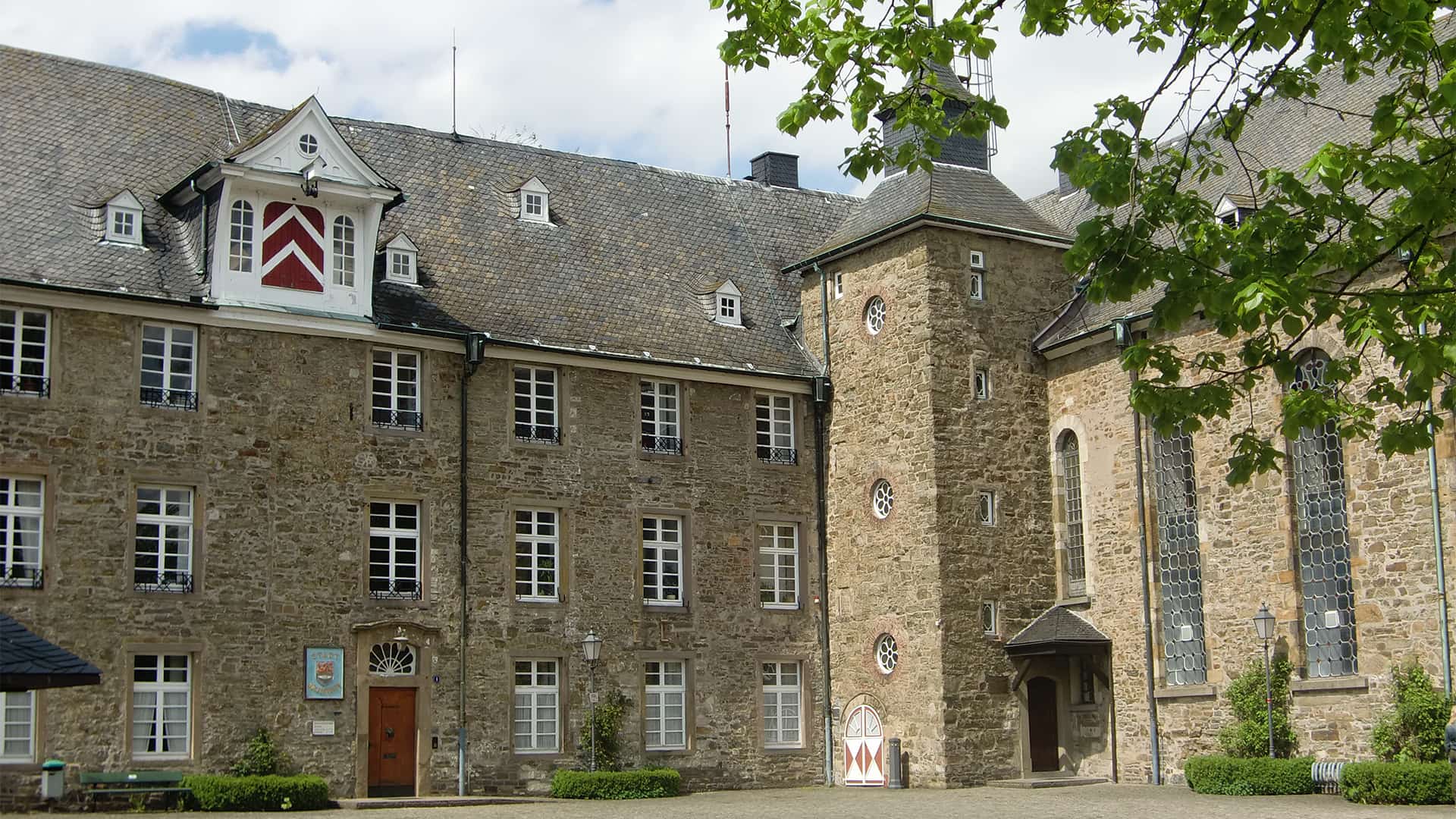 Steingebäude mit Turm, vielen Fenstern und Bäumen im Vordergrund.