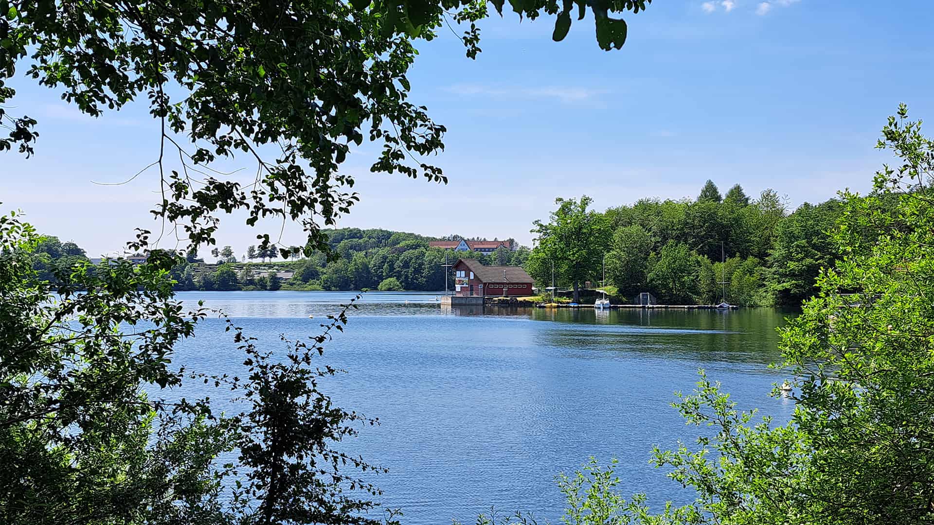 Blauer See, grüne Bäume, kleines Haus am Wasser, klarer Himmel.