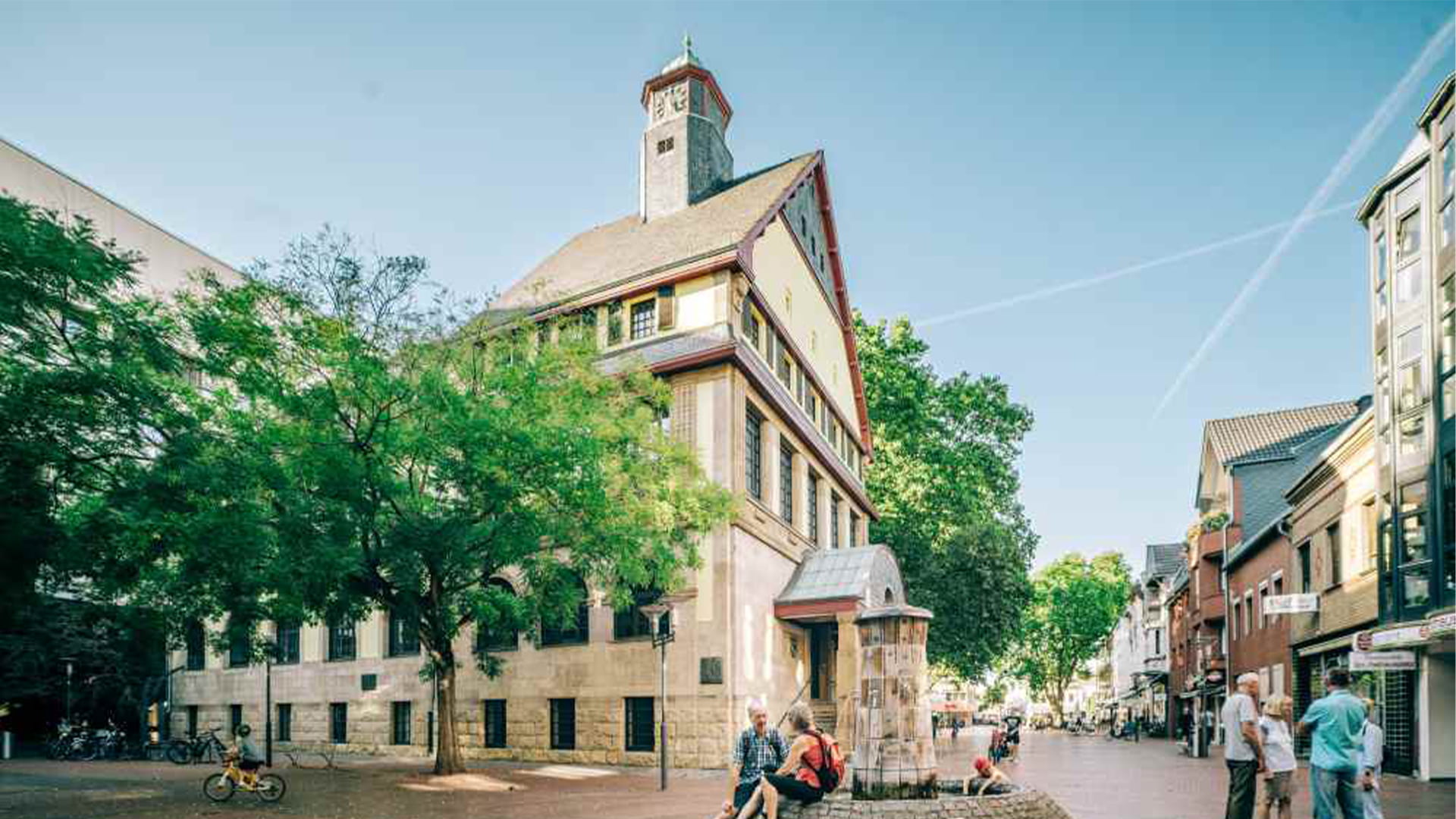 Sonniger Tag: Menschen am Brunnen vor historischem Gebäude.