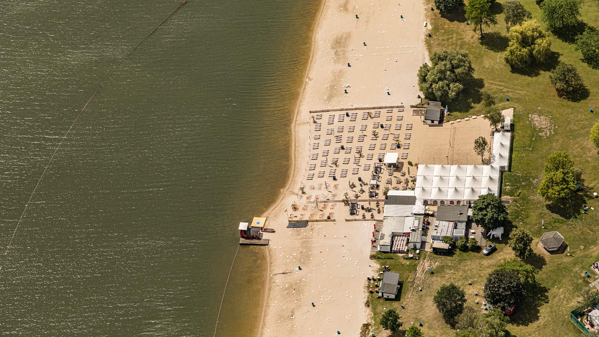 Strand mit Sonnenliegen, Schirmen, Gebäuden und Bäumen am Wasser.