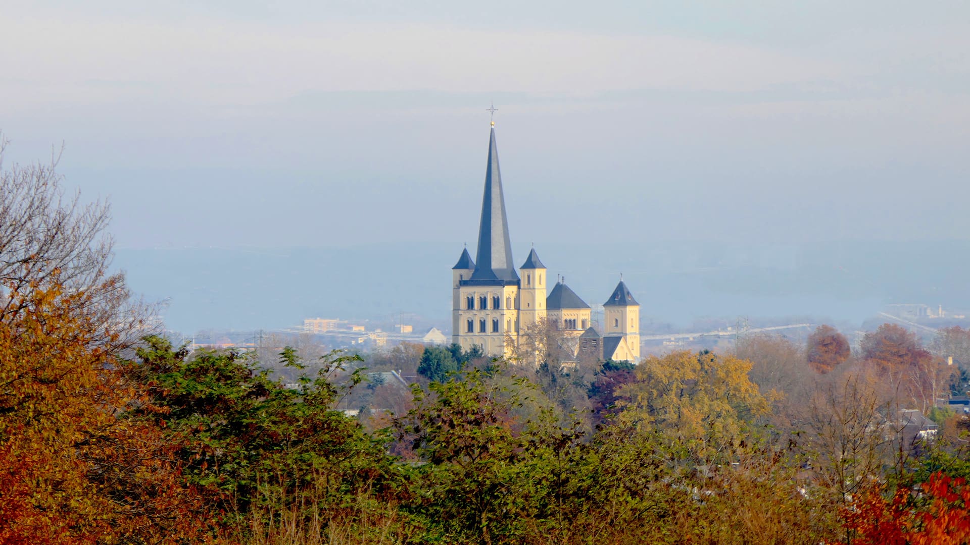 Die Kirchturmspitze überragt die herbstlichen Bäume unter einem dunstigen Himmel.