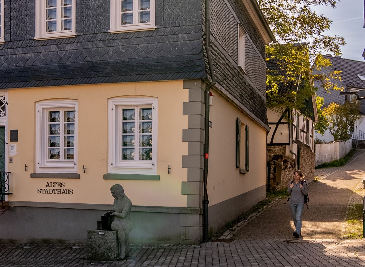 Frau geht an gelbem Gebäude, grauem Dach, Statue auf Kopfsteinpflaster vorbei.