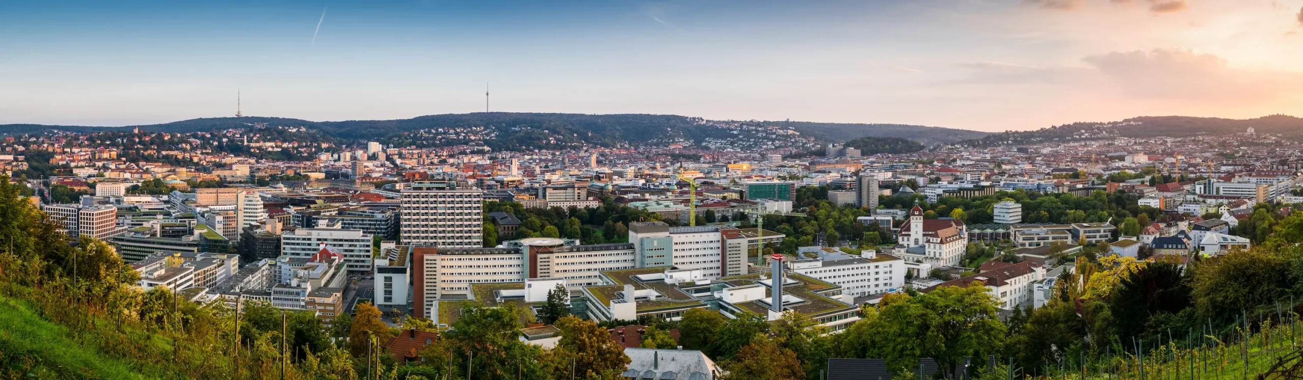 Stadt mit modernen Gebäuden, Hügeln und Grün im Sonnenuntergang.