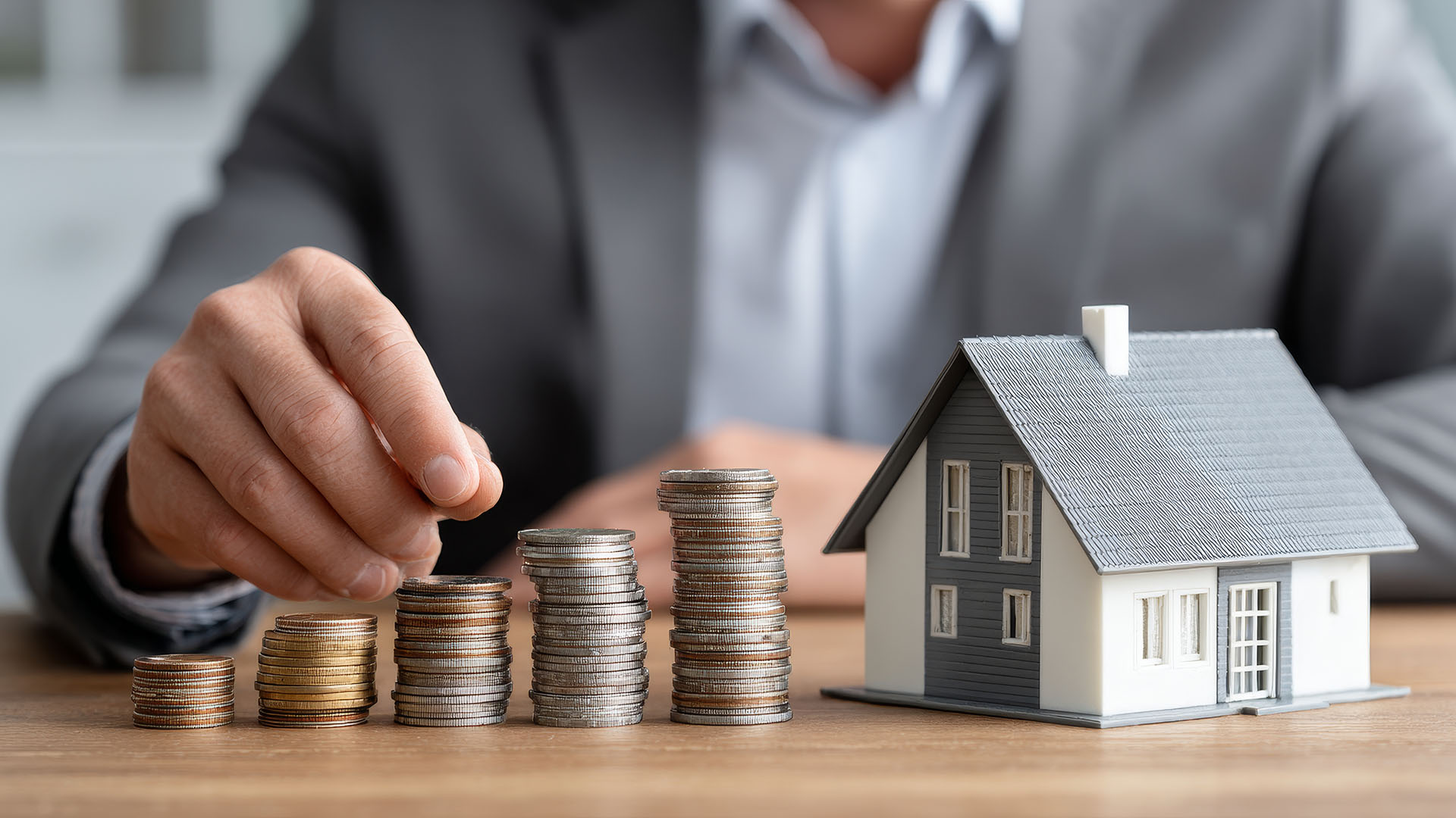 Hand stapelt Münzen neben einem Modellhaus.