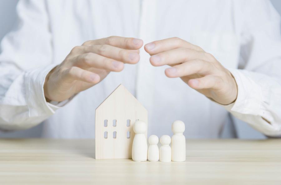 Mann hält schützend die Hände über ein Modellhaus mit Figuren