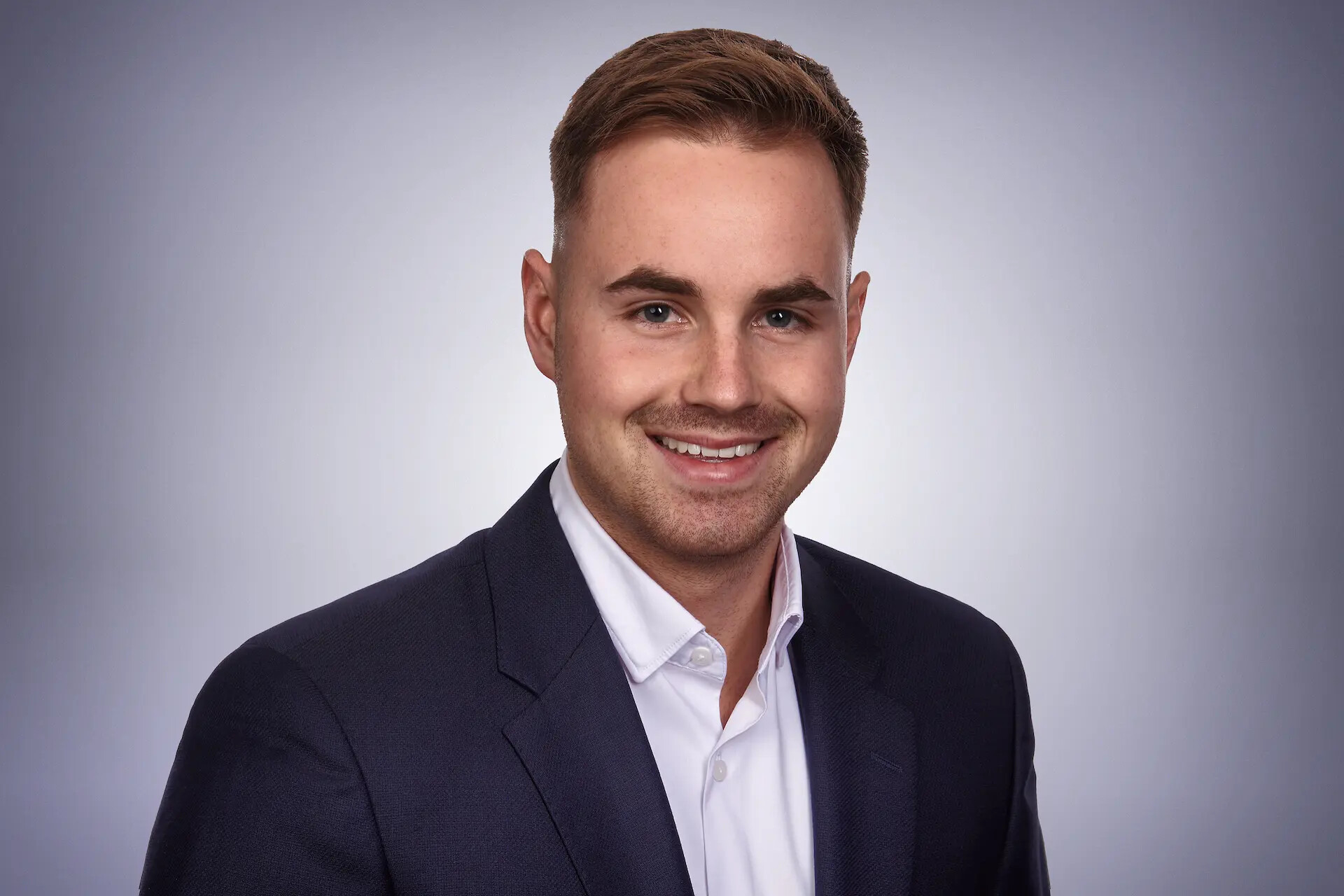 Benjamin Lambert in marineblauem Anzug und weißem Hemd vor grauem Hintergrund mit Farbverlauf.