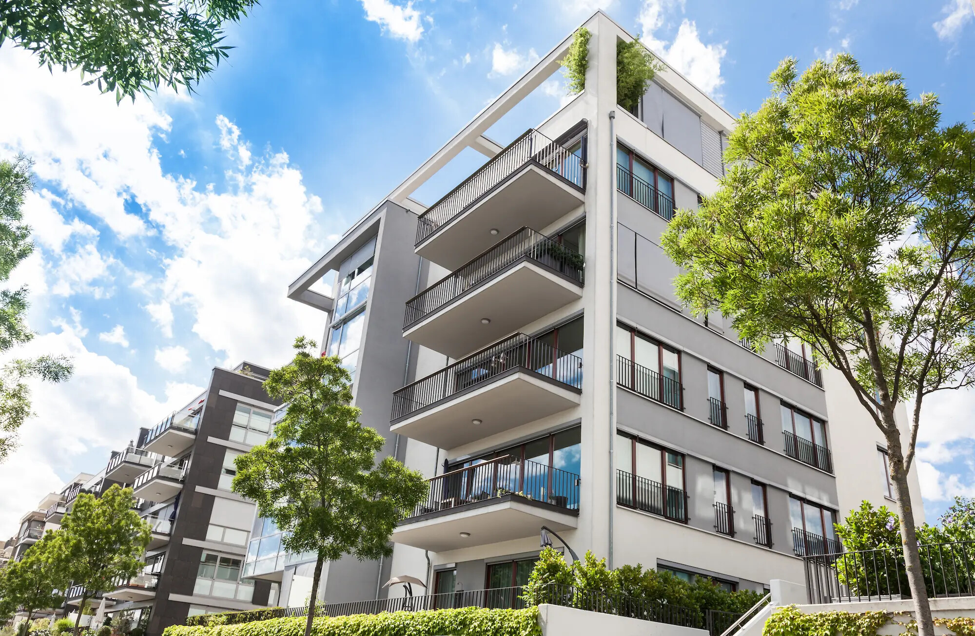 Mehrfamilienhaus mit Balkonen, Bäumen und blauem Himmel.
