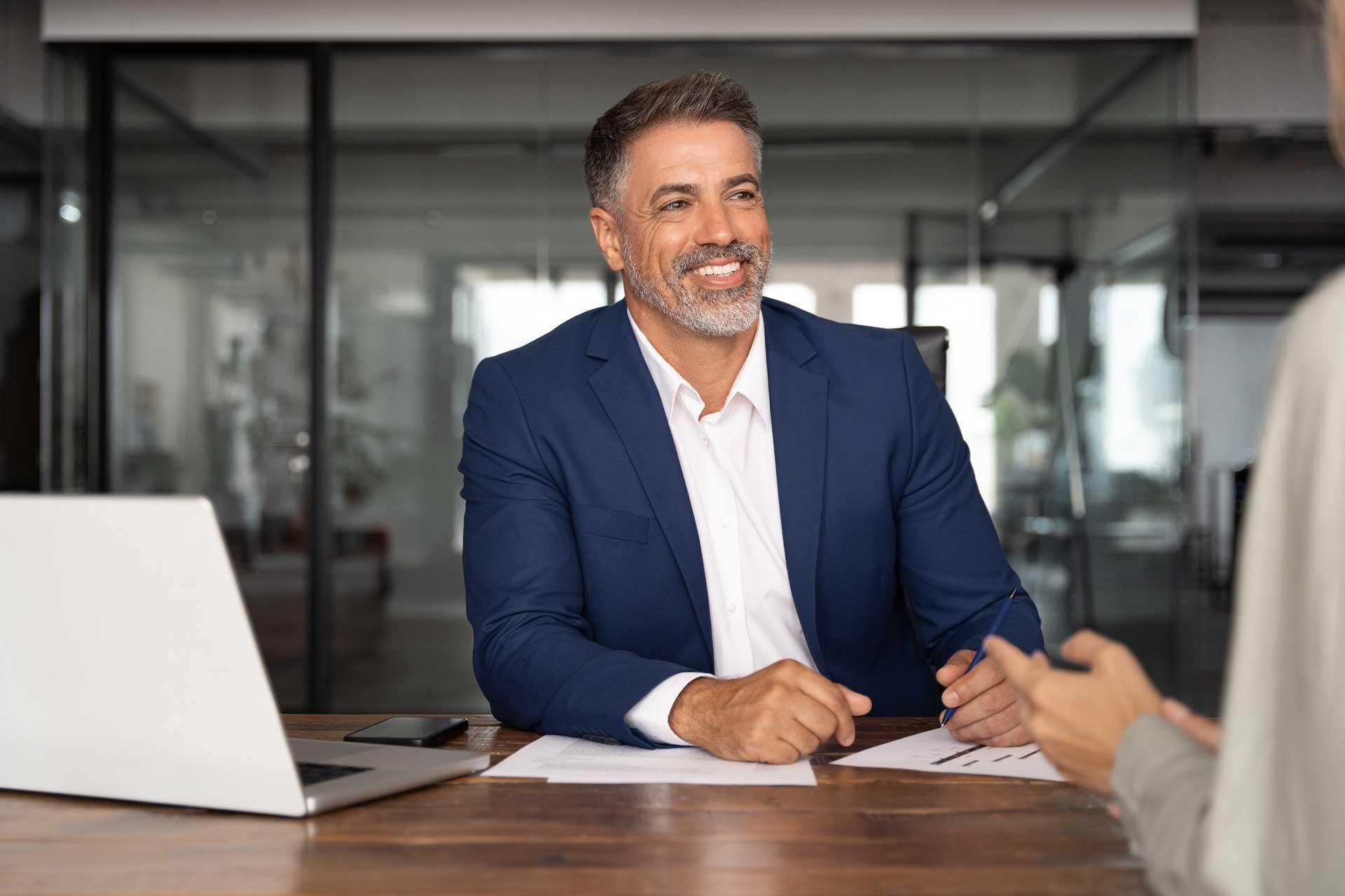 Mann im blauen Anzug am Schreibtisch mit Laptop, spricht mit Person.