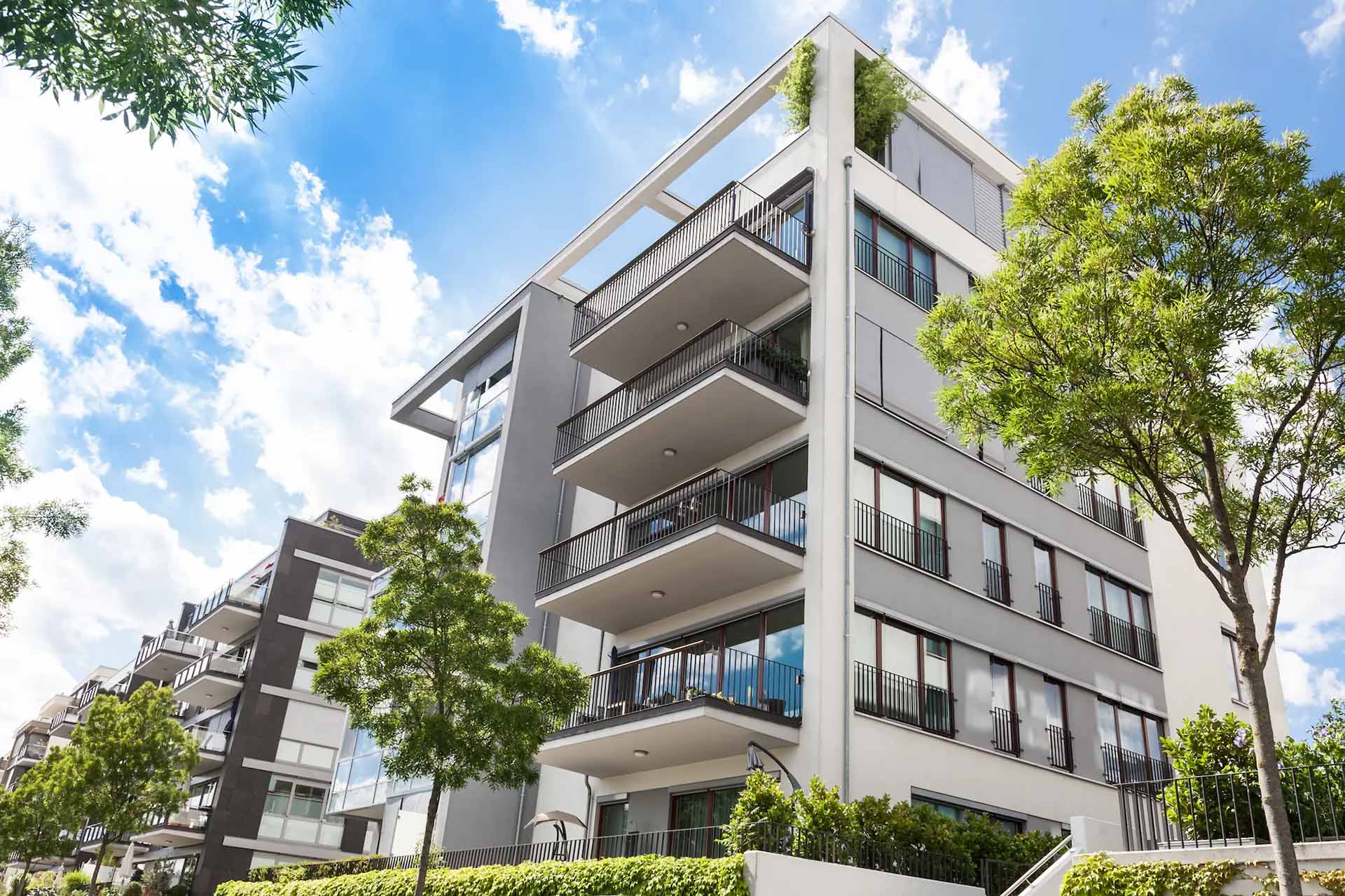 Mehrfamilienhaus mit Balkonen, Bäumen und blauem Himmel.