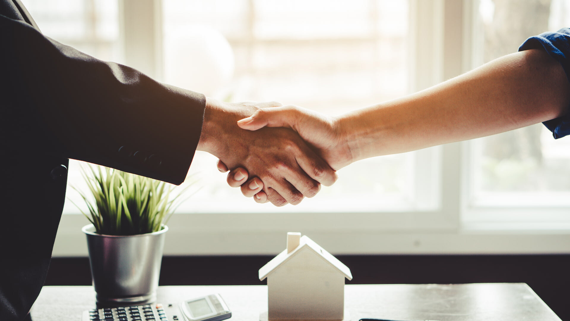 Händedruck über Schreibtisch mit Hausmodell und Pflanze.
