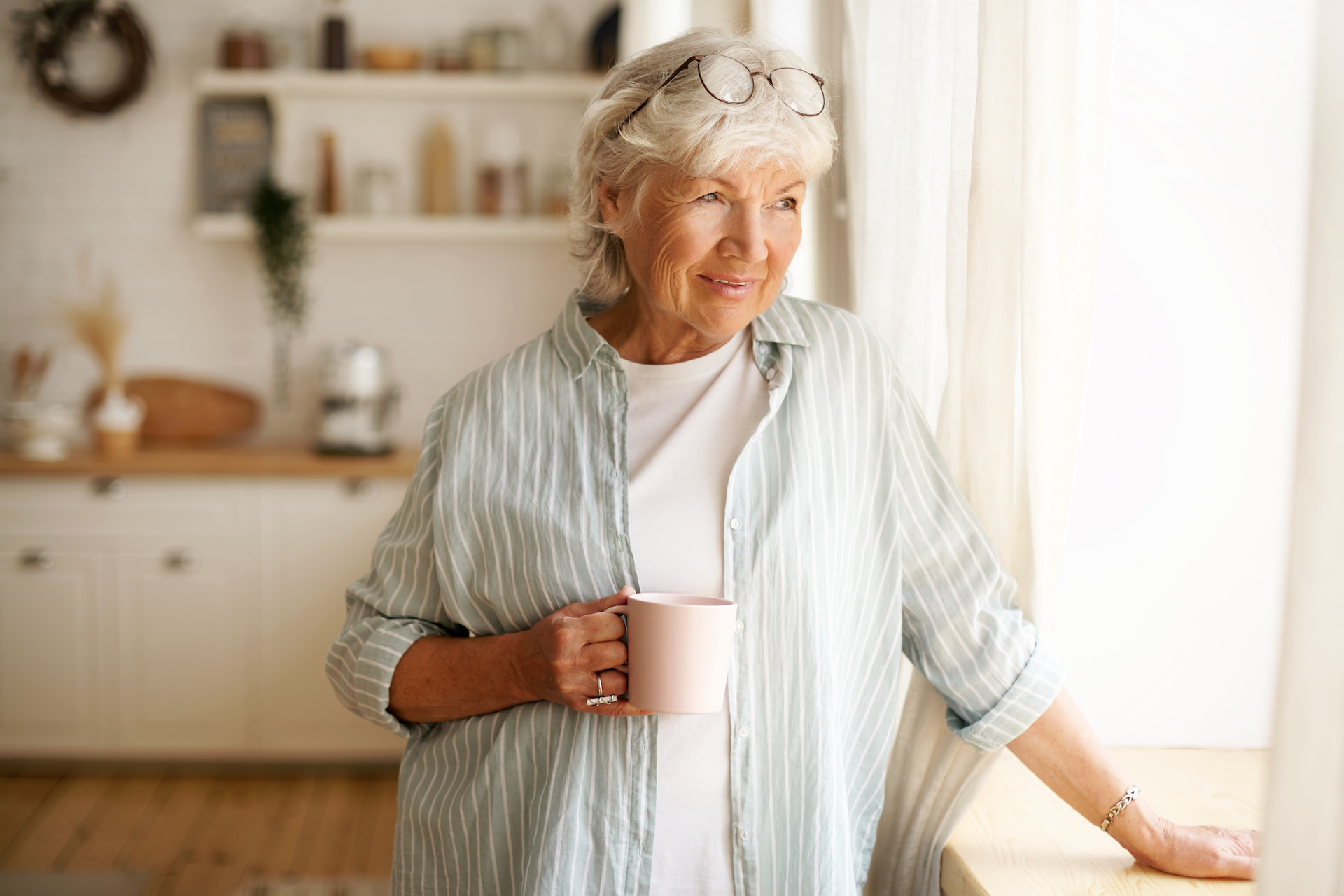 Ältere Frau mit grauen Haaren hält Tasse und lächelt am Fenster.