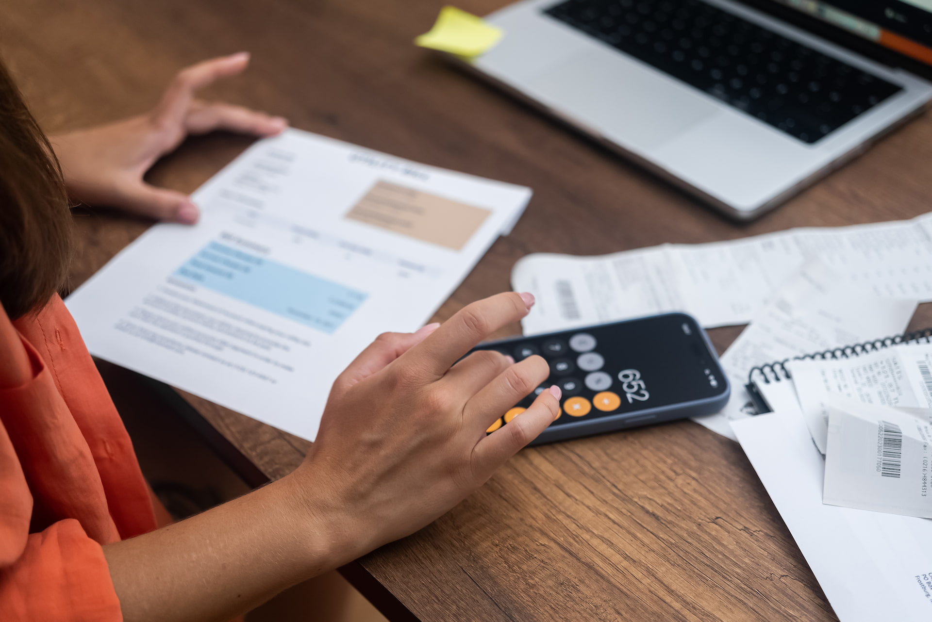 Person mit Rechner, Finanzdokumenten und Laptop am Schreibtisch.