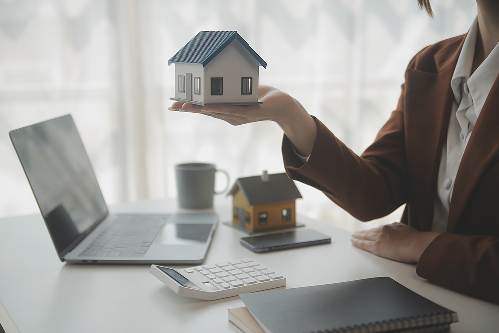 Person im Anzug hält Modellhaus am Schreibtisch mit Laptop.