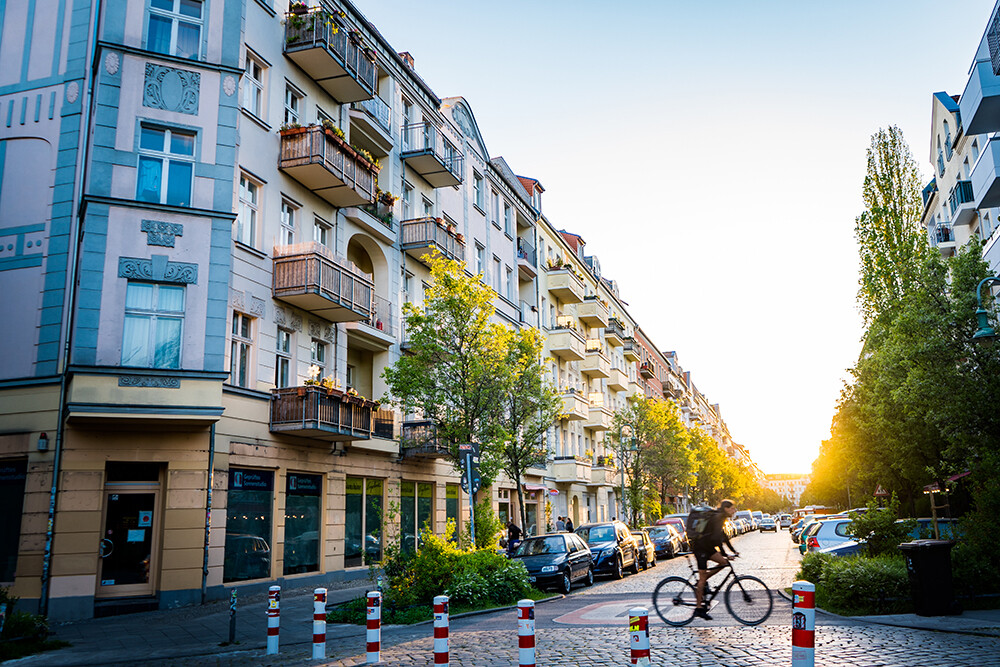 Person auf Fahrrad fährt sonnige Straße mit Autos und bunten Häusern.