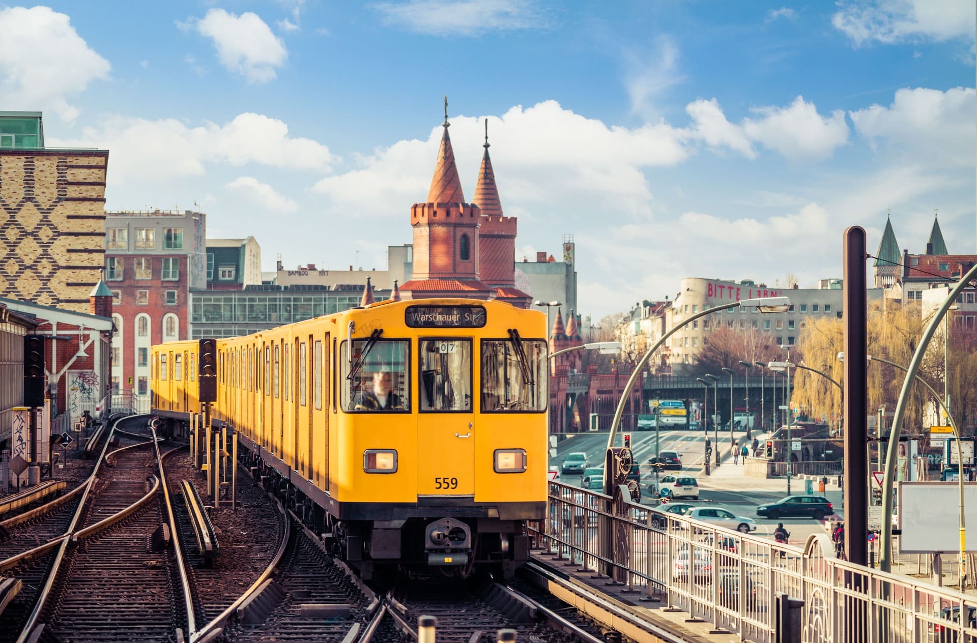 Gelber Zug fährt auf Hochbahn vor historischen Stadtgebäuden.