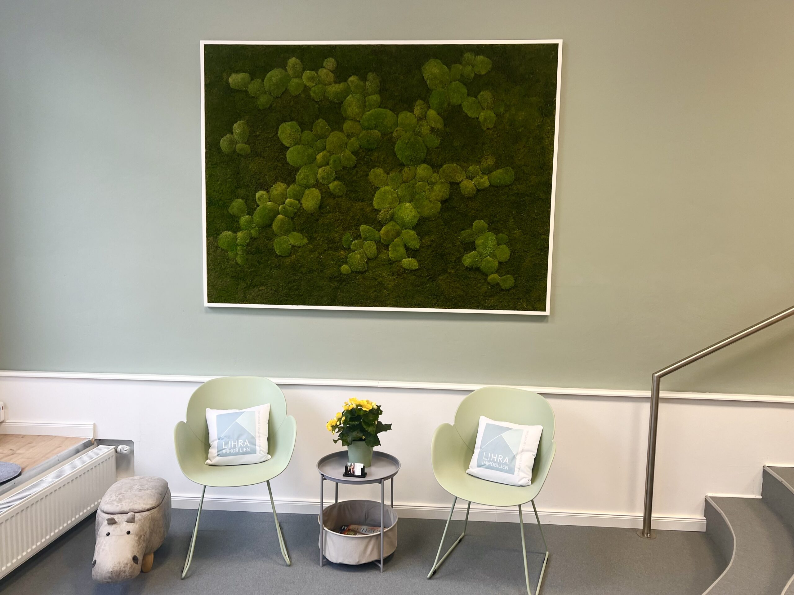 Zwei grüne Stühle, runder Tisch mit Pflanze, Moosbild.