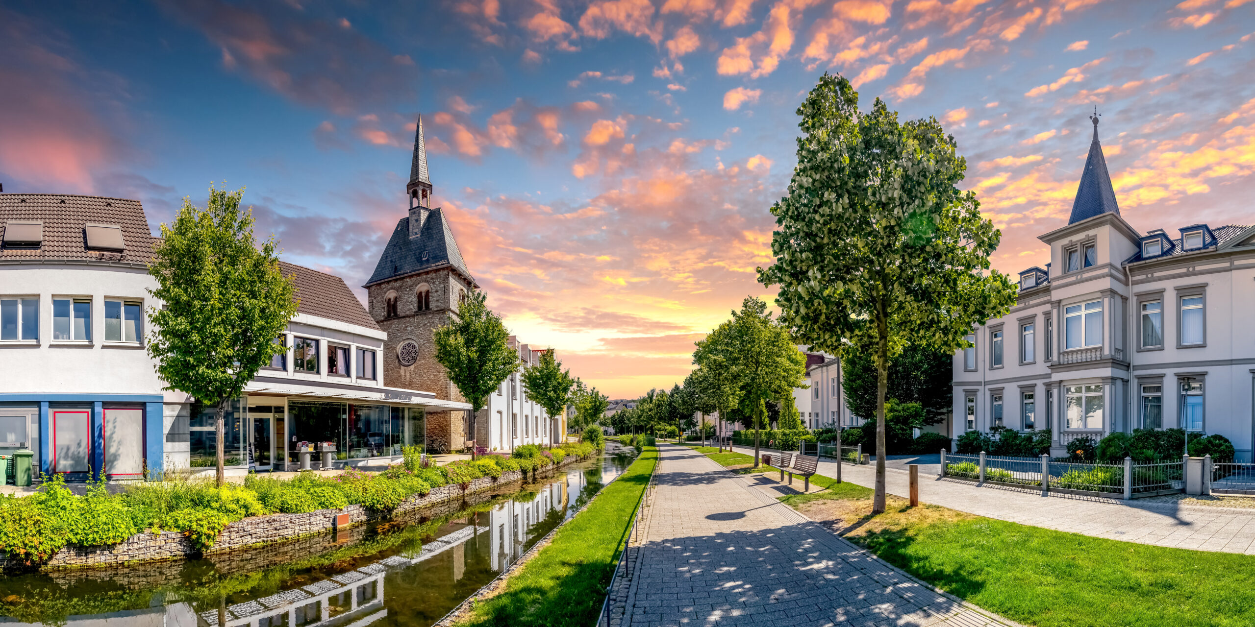 Malerischer Weg mit Bäumen, Gebäuden und Kirche bei Sonnenuntergang.