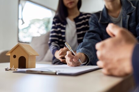 Paar unterschreibt Vertrag, daneben ein kleines Modellhaus aus Holz