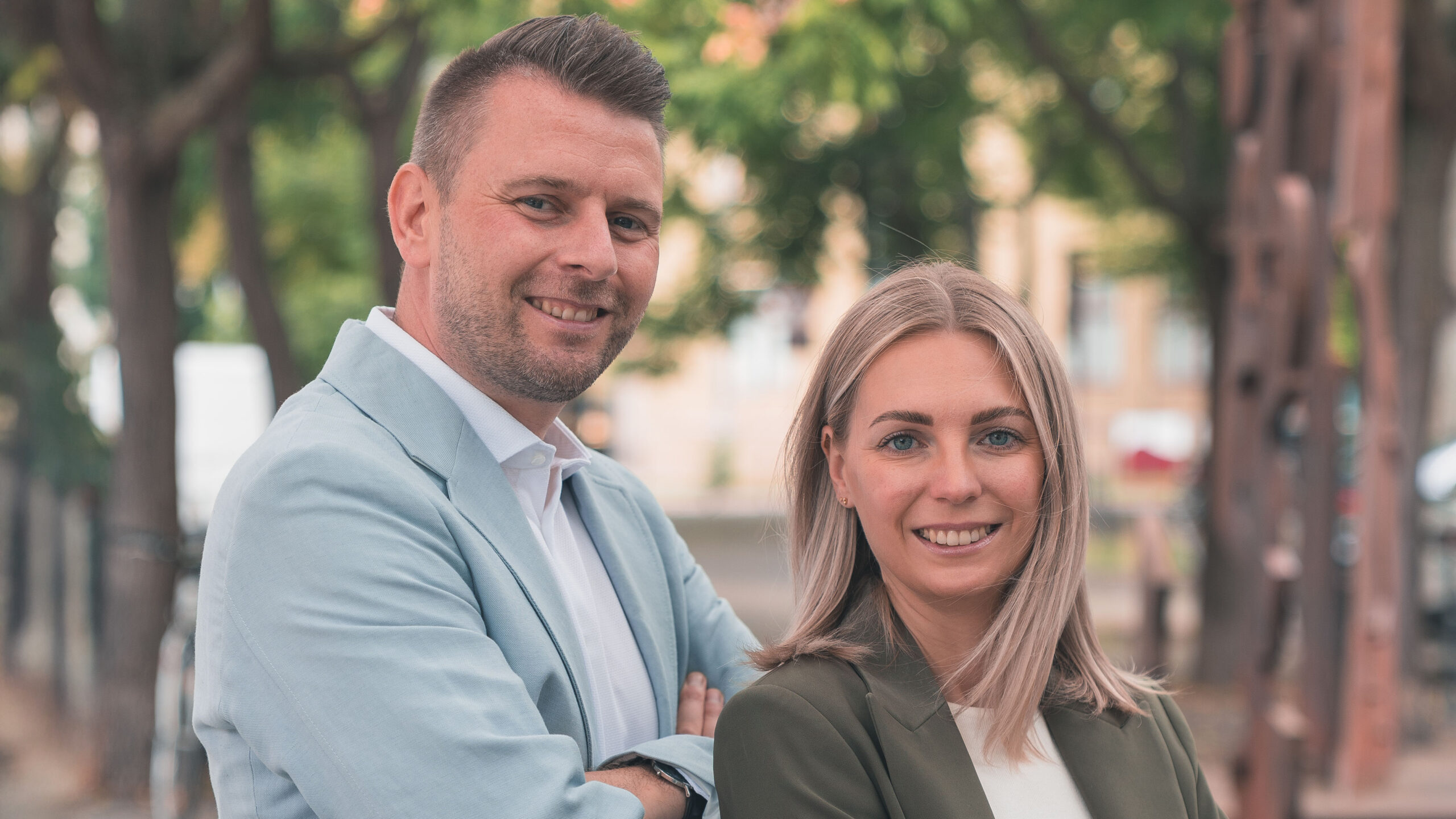 Christopher und Marika Oehlschlegel im Business-Look lächeln draußen vor Bäumen.