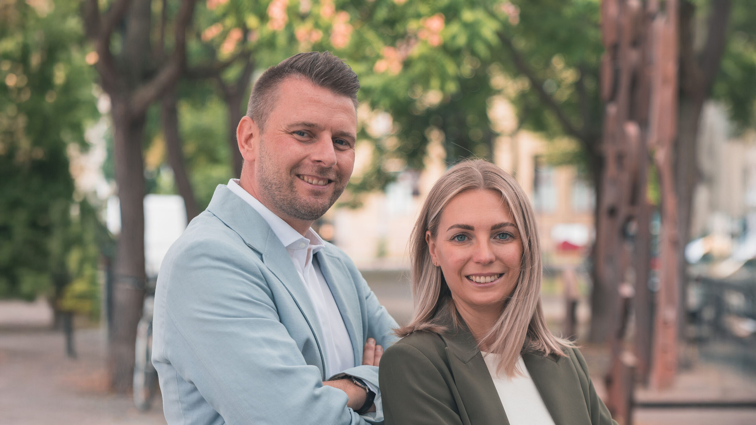 Christopher und Marika Oehlschlegel im Business-Look lächeln draußen vor Bäumen.