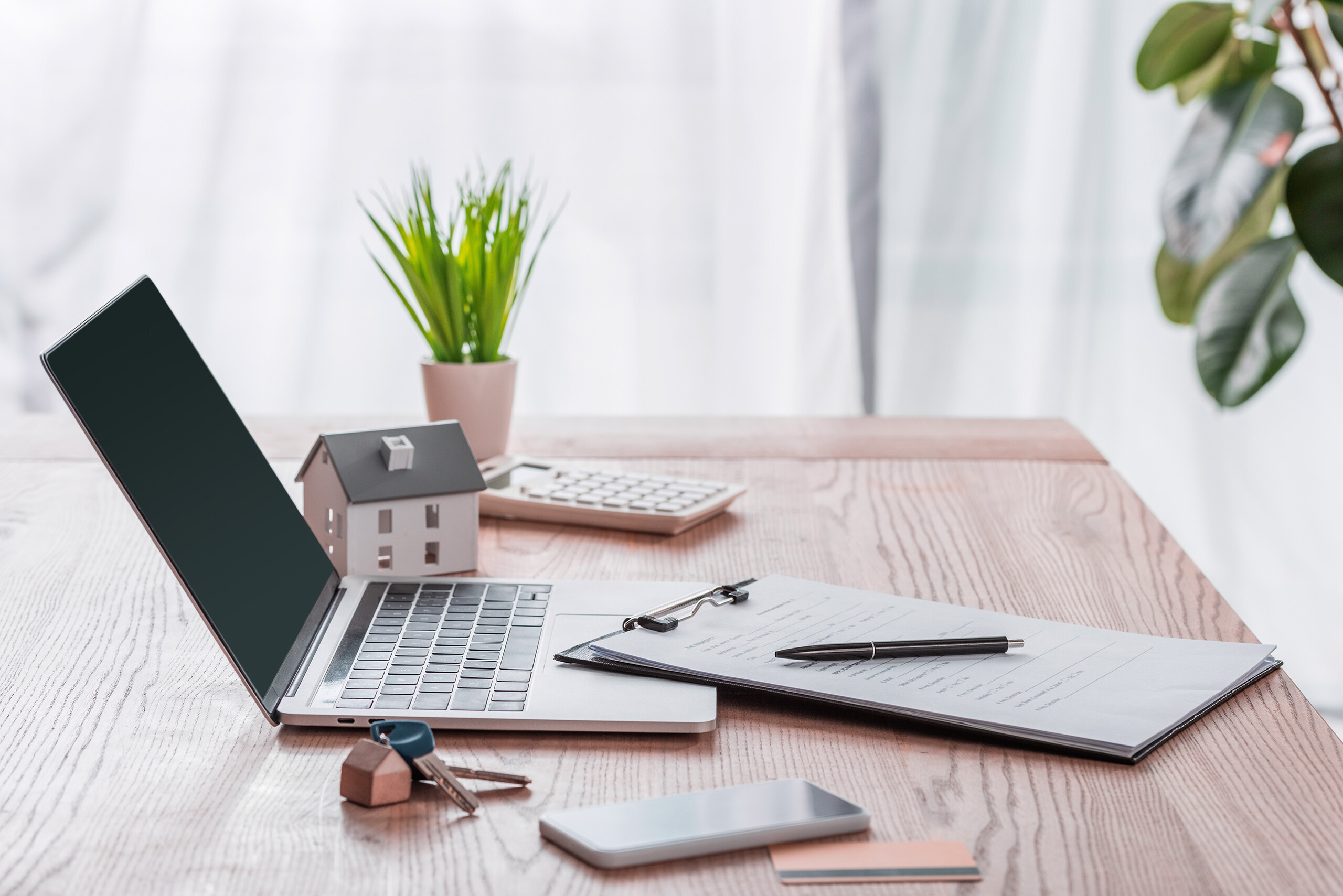 Laptop, Hausmodell, Schlüssel und Klemmbrett auf Holzschreibtisch.