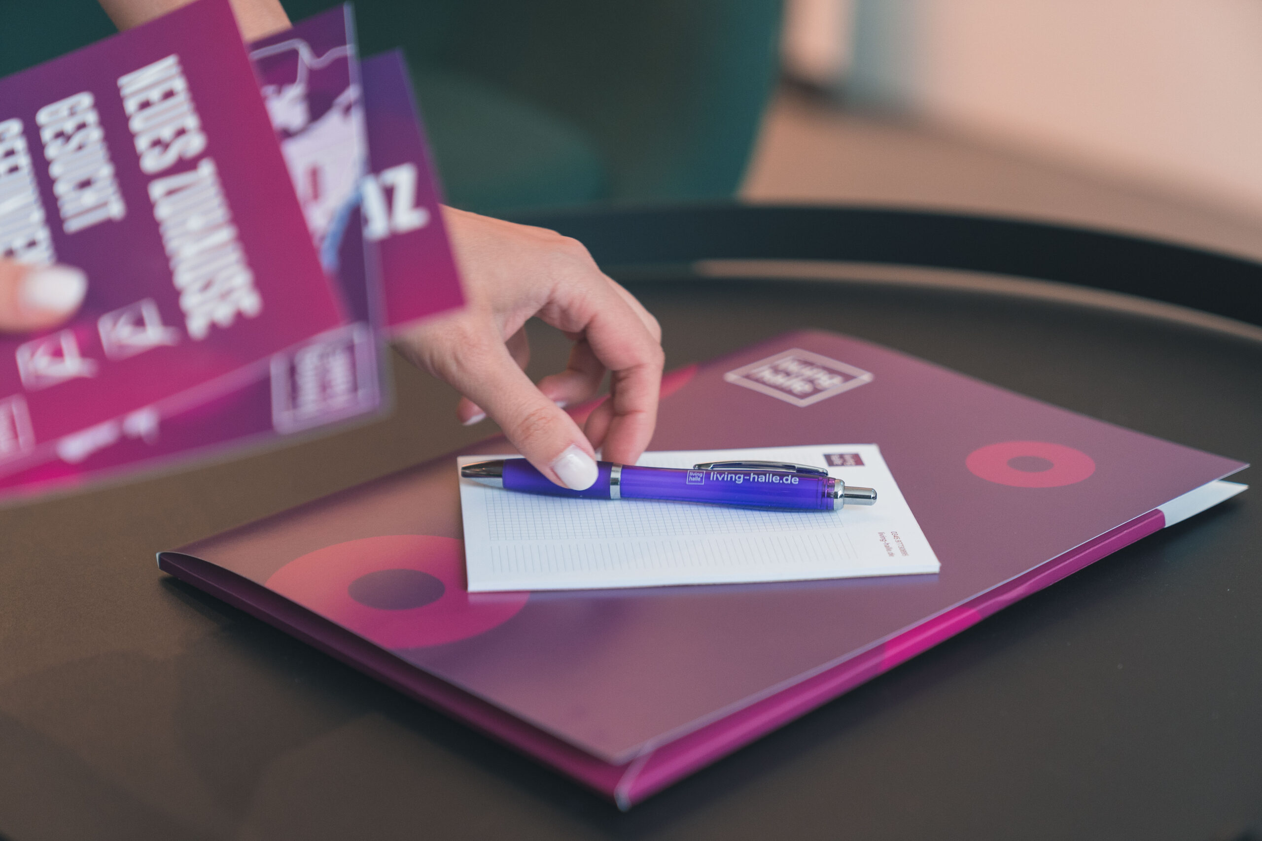 Hand nimmt lila Broschüre neben Notizbuch und Stift auf.