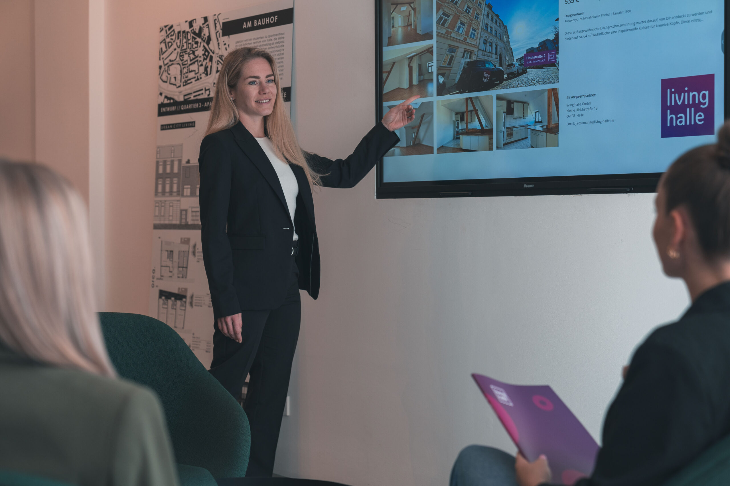 Frau im Anzug zeigt zwei Frauen am Bildschirm Immobiliendetails.