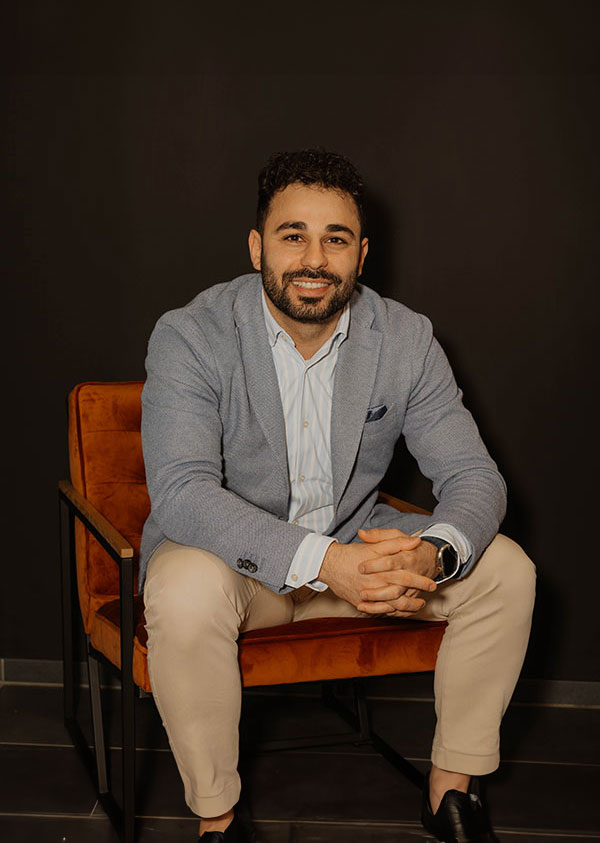 Mann mit Bart in hellem Blazer lächelt auf orangem Stuhl.