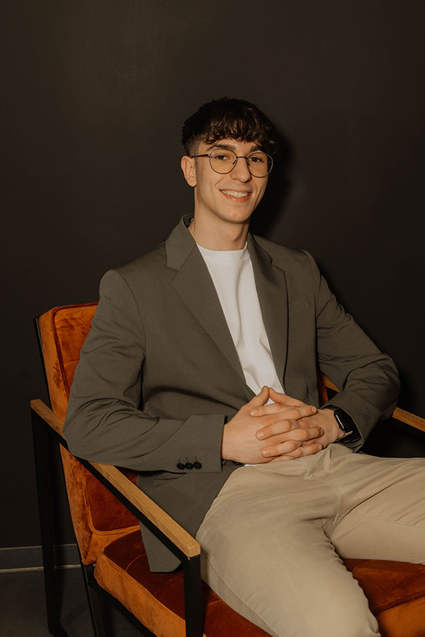 Junger Mann mit Brille lächelt auf orangem Stuhl vor dunklem Hintergrund.