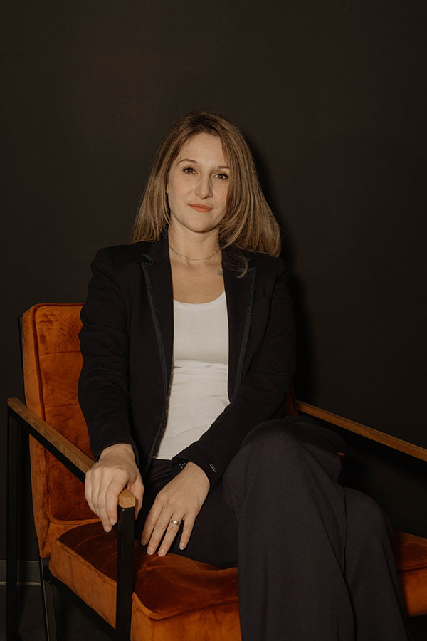 Frau in dunklem Anzug auf orangefarbenem Stuhl, dunkler Hintergrund, Blick in die Kamera.