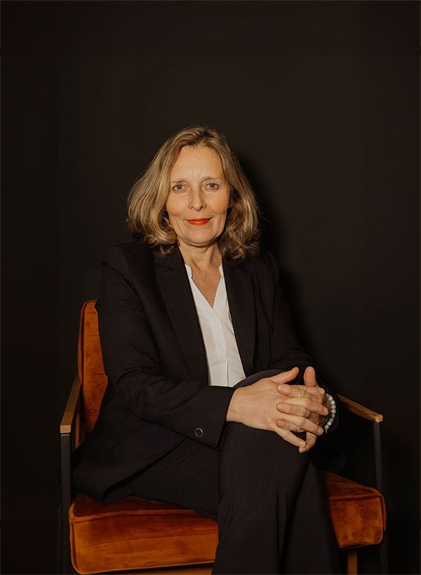 Frau in schwarzem Anzug auf orangefarbenem Stuhl, dunkler Hintergrund, Hände gefaltet.