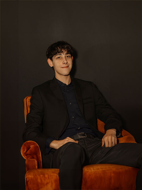 Mann im schwarzen Anzug sitzt selbstbewusst im orangen Sessel.