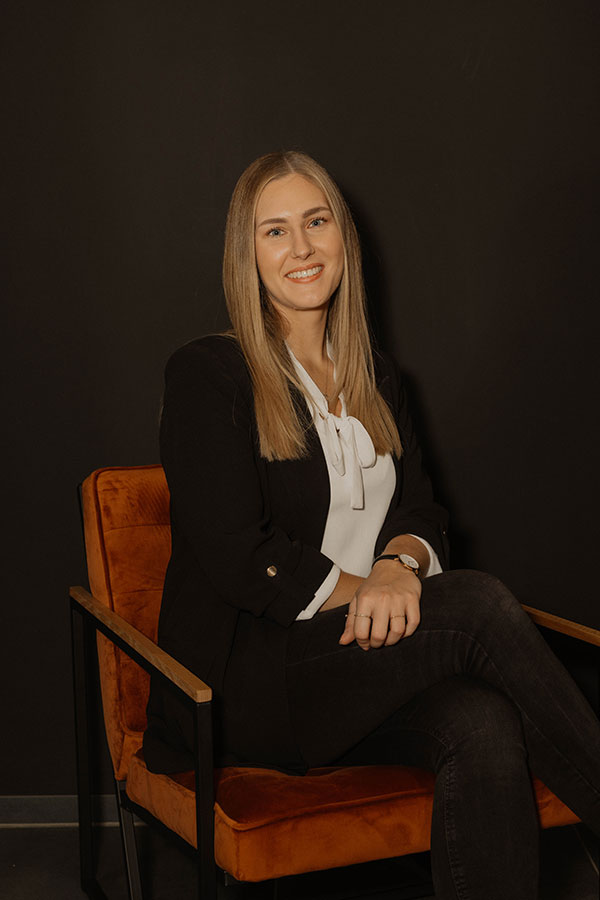 Frau mit glattem Haar lächelt auf orangem Stuhl vor dunklem Hintergrund.