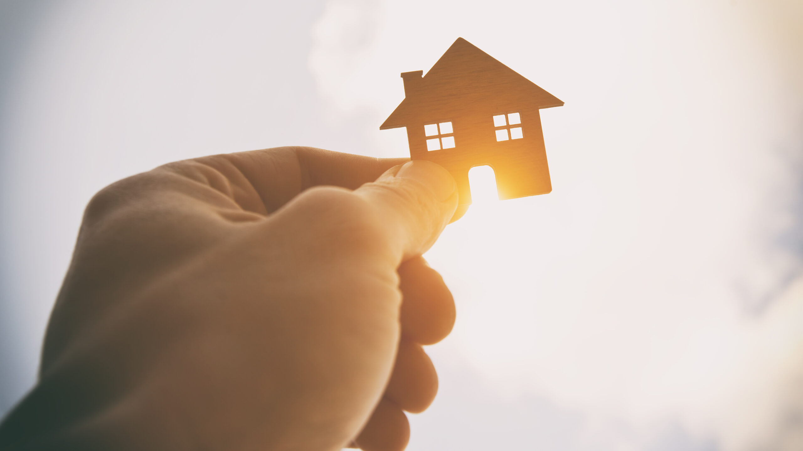 Hand hält kleines Haus vor hellem, sonnigem Himmel.