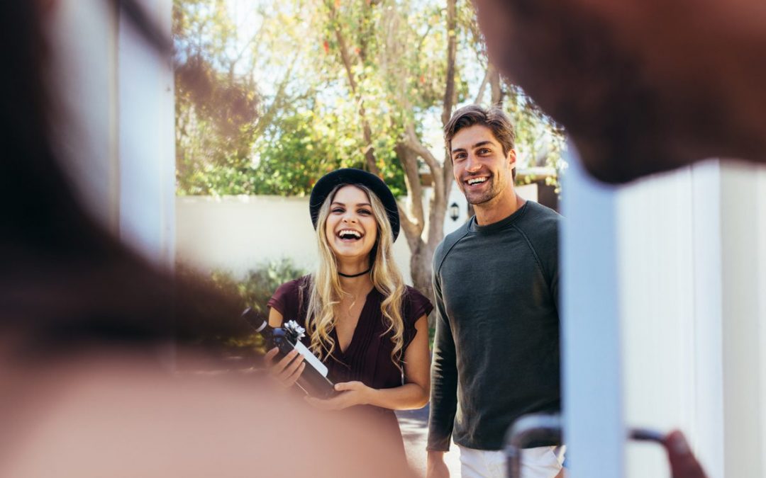 Frau und Mann draußen, begrüßen jemanden an einer Tür, Flasche in der Hand.