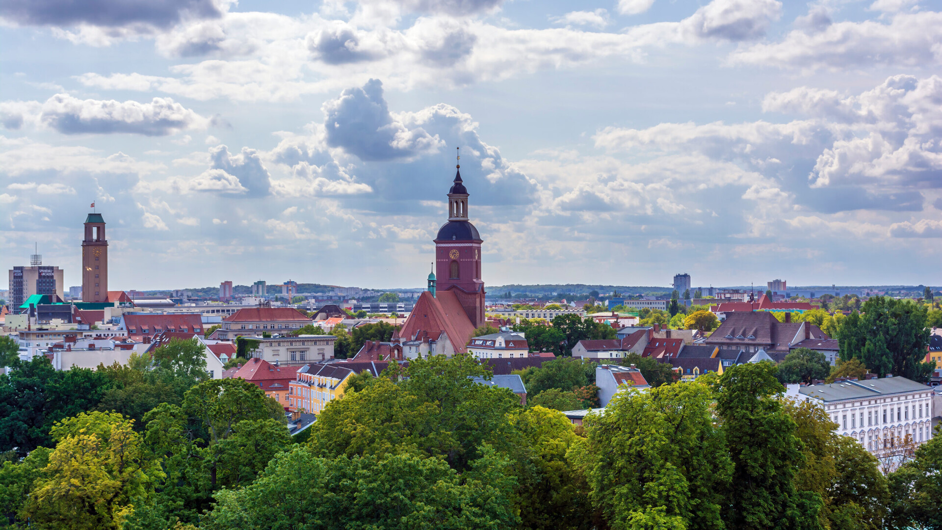 Stadt mit Kirchtürmen, roten Dächern und Bäumen unter Wolken.