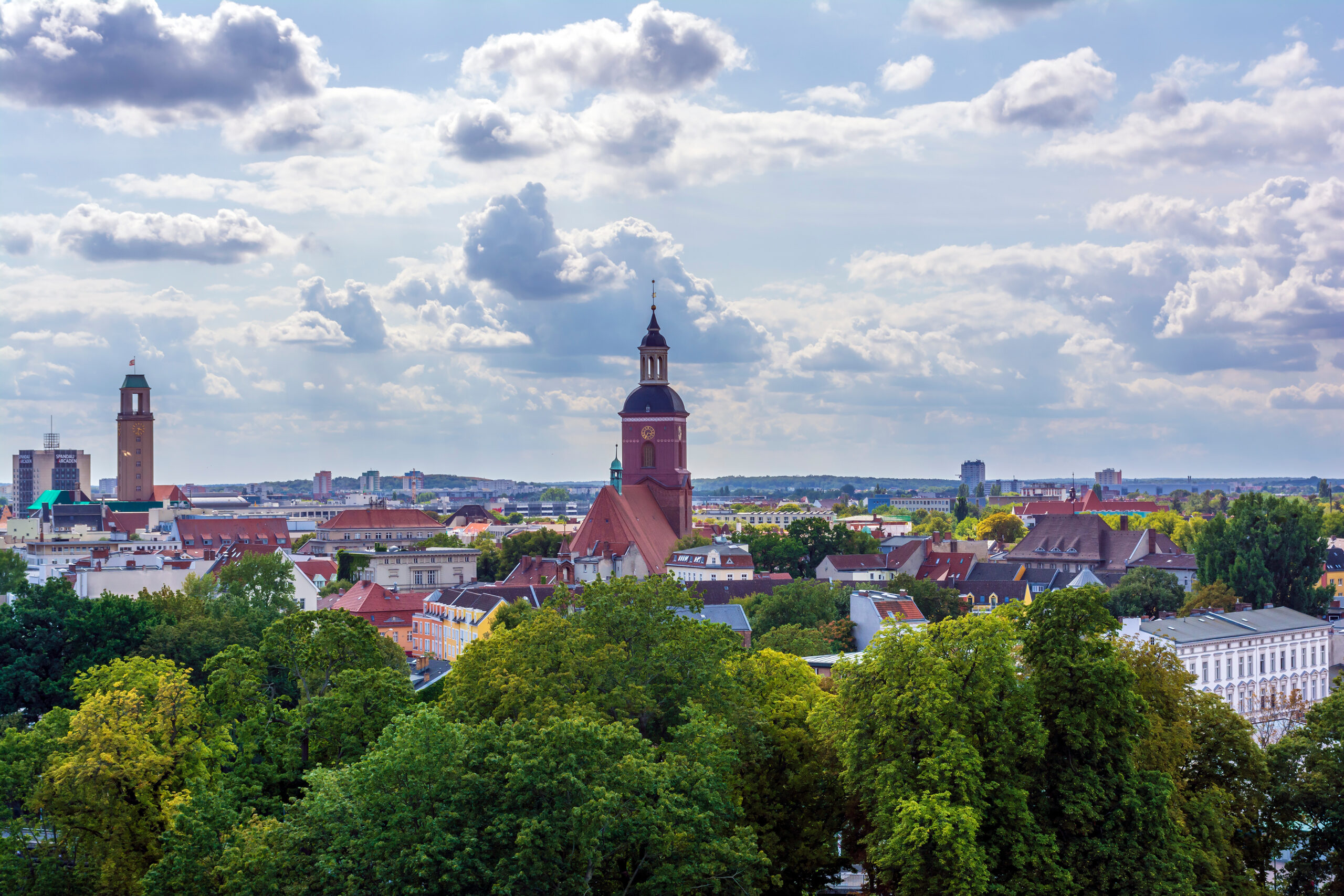 Stadt mit Kirchtürmen, roten Dächern und Bäumen unter Wolken.