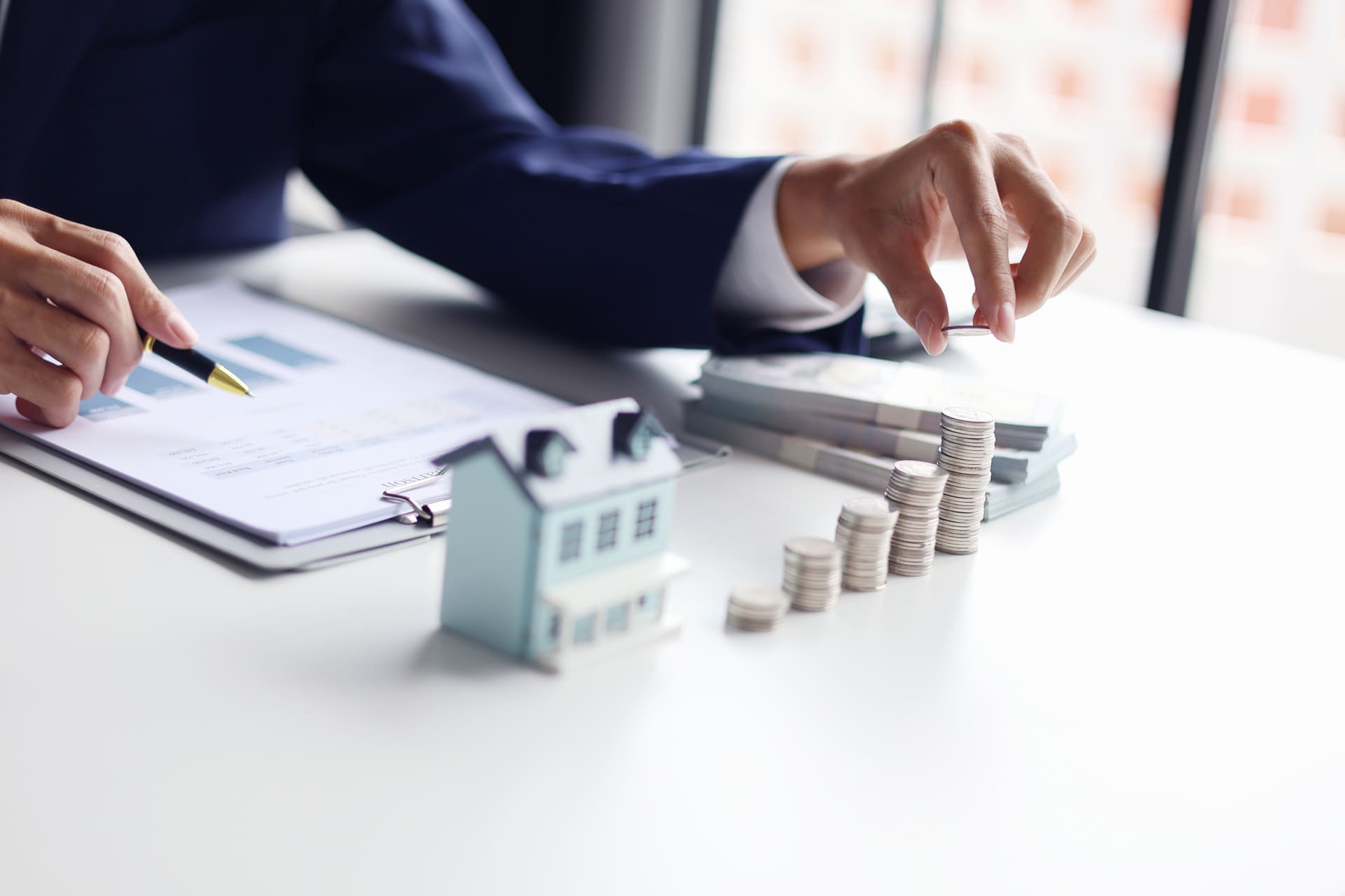 Hand hält Münze neben Miniaturhaus und Vertrag auf Tisch.